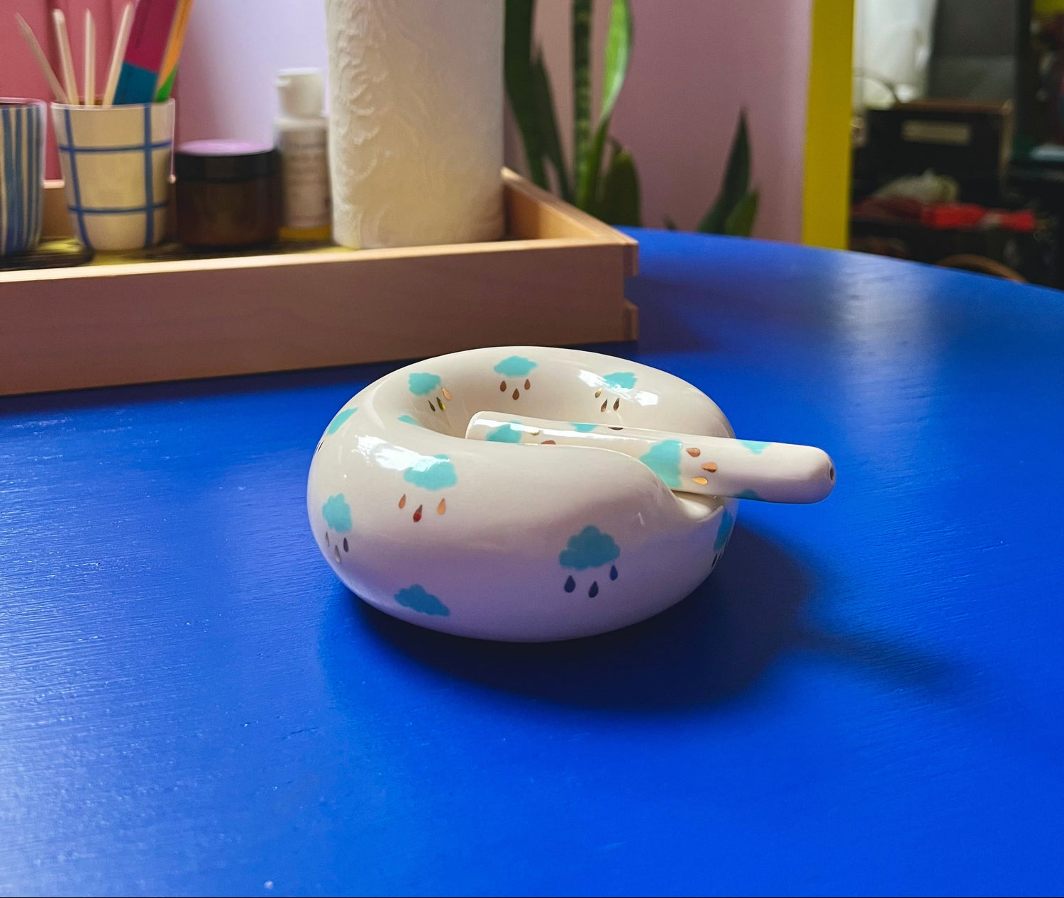 Small white container with cloud designs on a blue surface, with a pink wall and green plant in the background.
